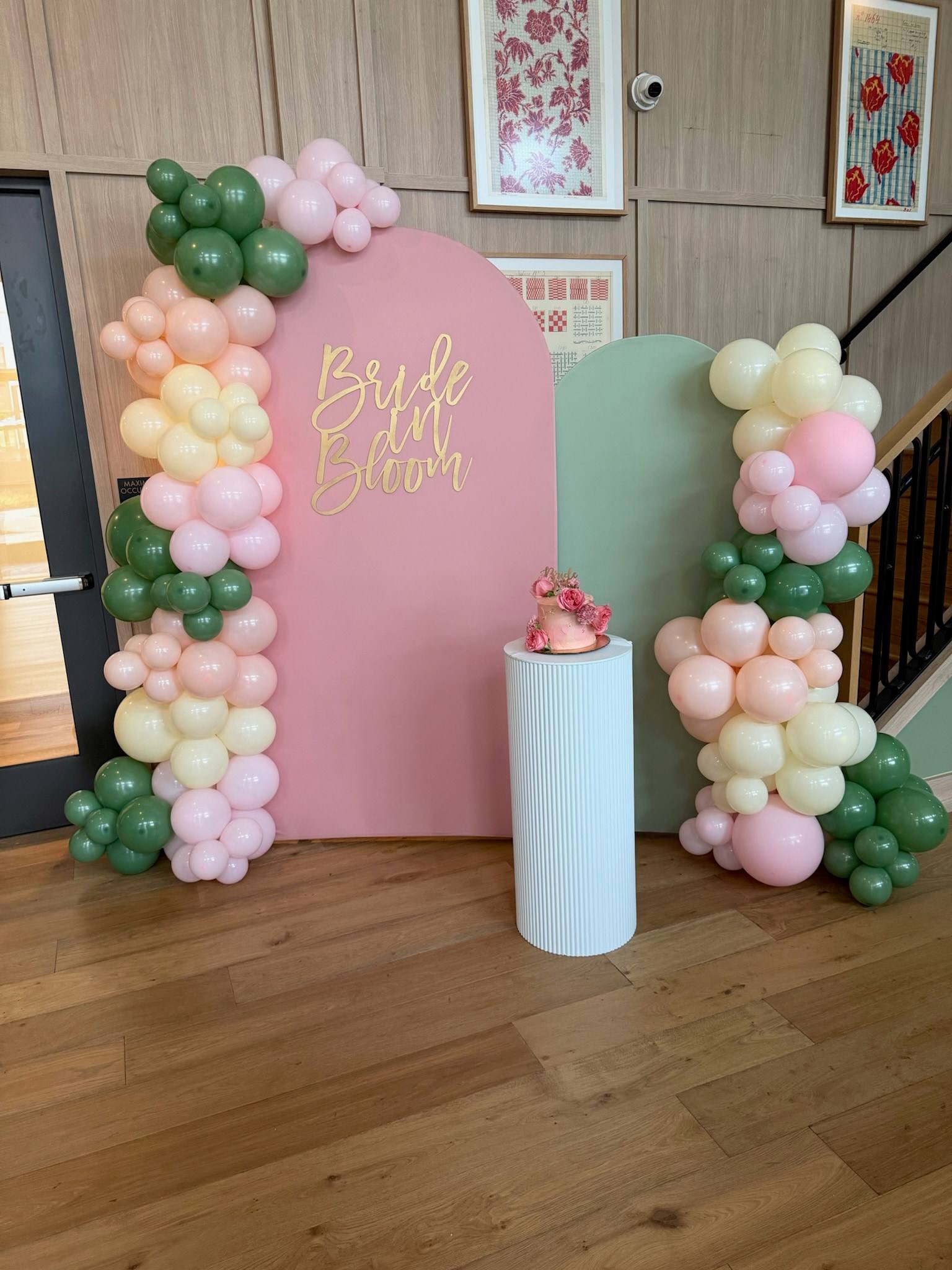 A pink arch backdrop lettered Bride in Bloom in gold, framed by pink, sage and cream balloon garlands with a white pedestal, indoors
