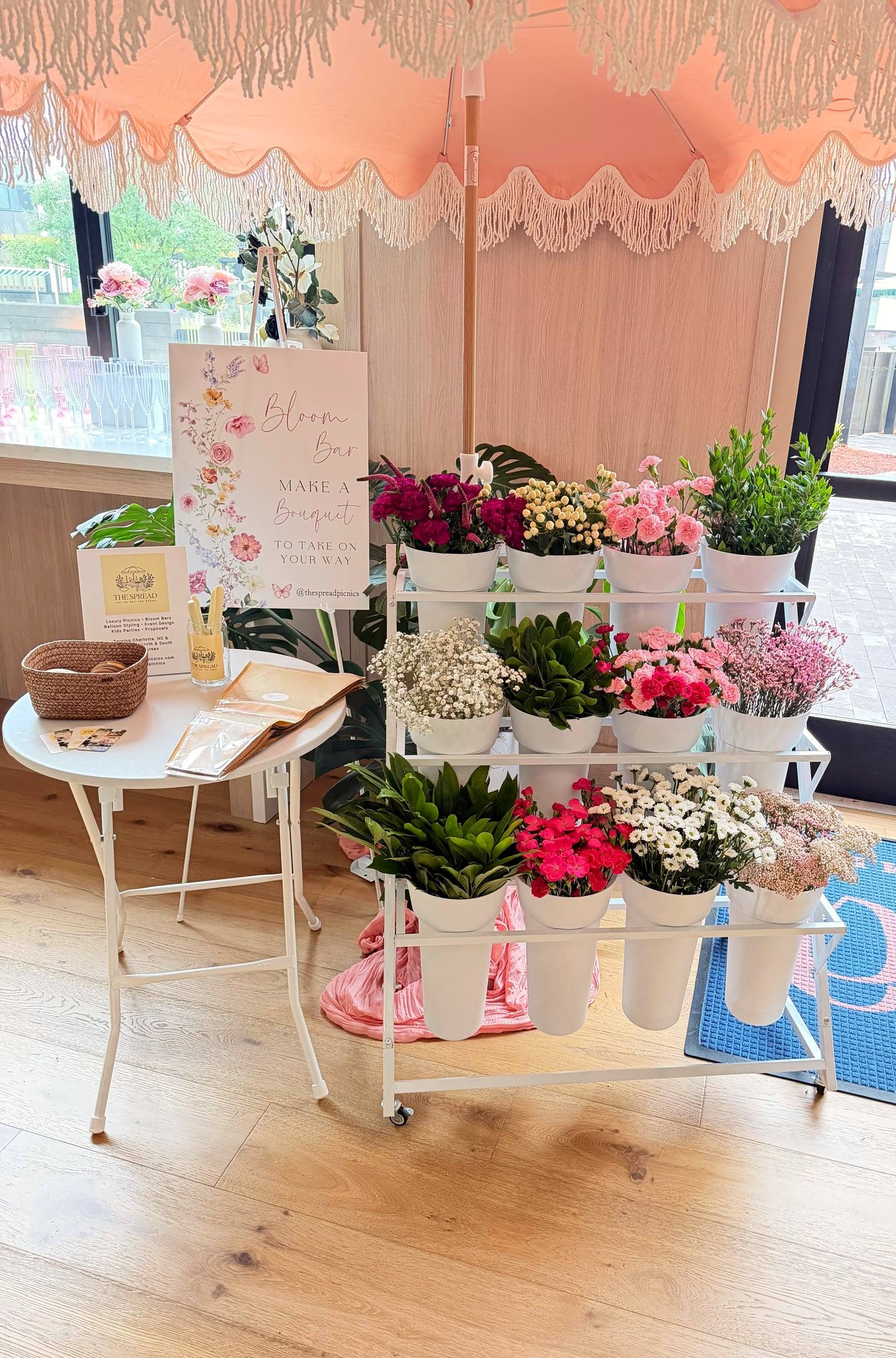 The white Bloom Bar cart under a blush fringed umbrella, three tiers of fresh stems in white buckets, with a Make a Bouquet sign and a side table of wrapping sleeves