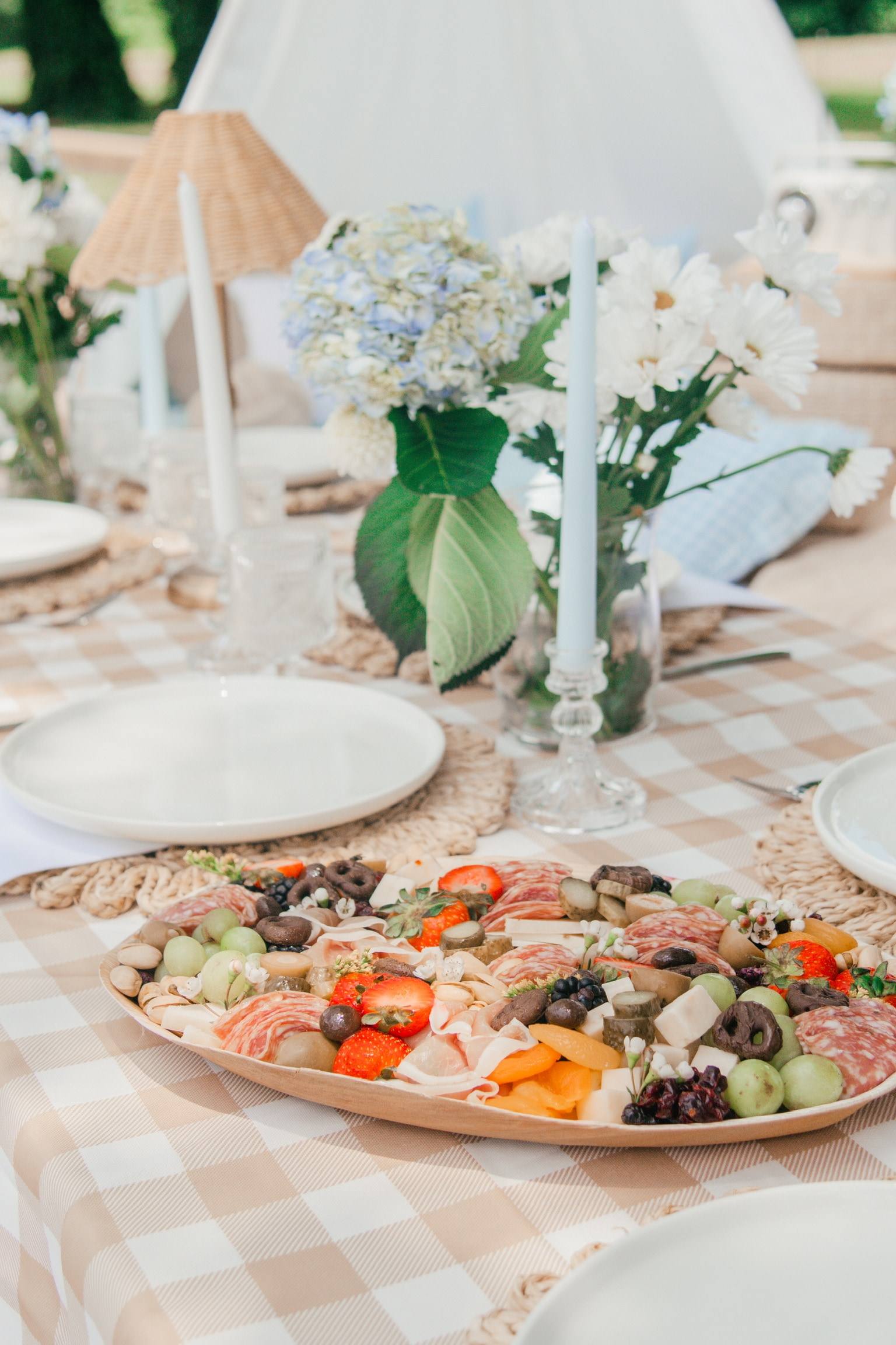 A charcuterie board of cured meats, cheese, strawberries and grapes on a gingham table, with a rattan lamp and a teepee behind