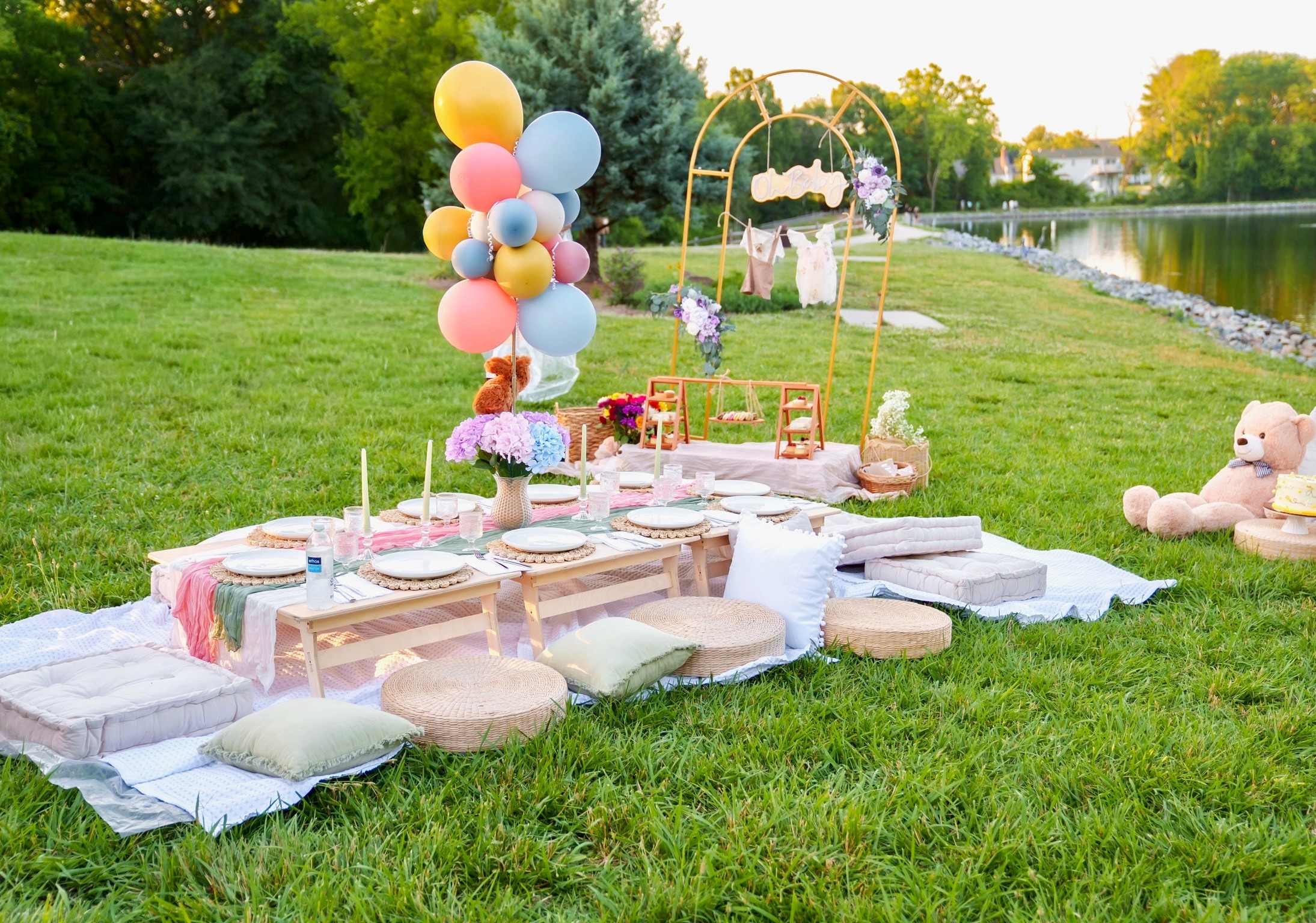 A baby shower picnic by a lake: a long low table with pastel linens, a pastel balloon bunch, a gold arch hung with baby clothes and a teddy bear
