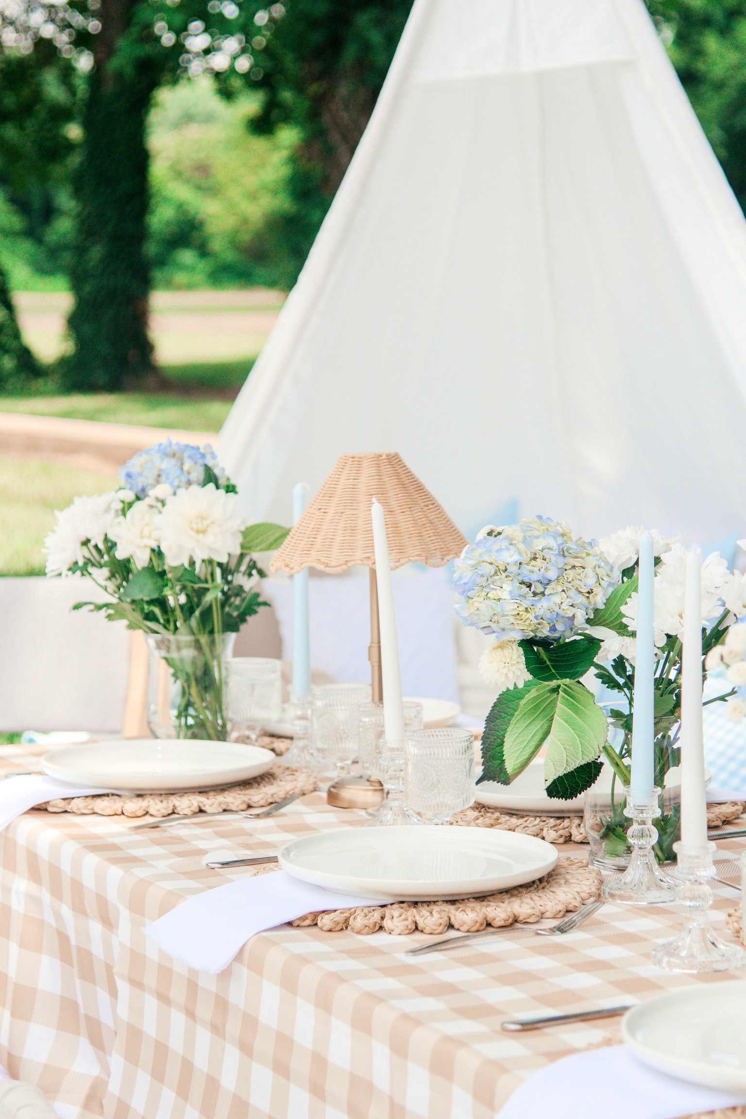 Blue hydrangeas and a rattan lamp on a gingham table, a white teepee behind