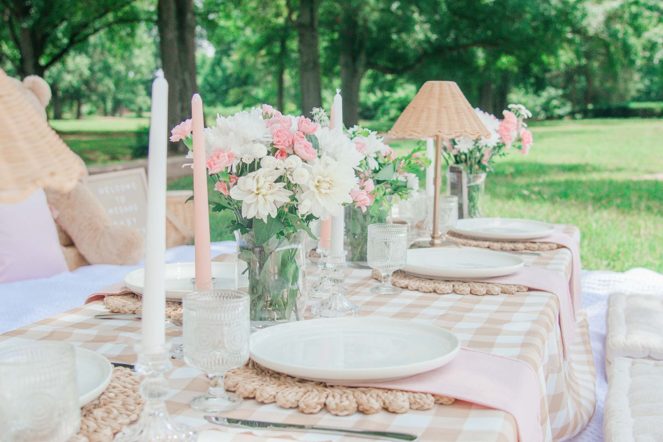 A gingham picnic table with pink flowers, a rattan lamp and tall candles