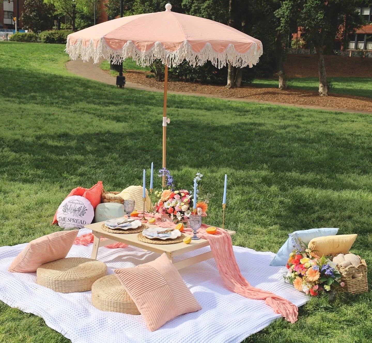 A low picnic table under a peach fringed umbrella, set with candles and flowers, with floor cushions and pillows on a white blanket in a park