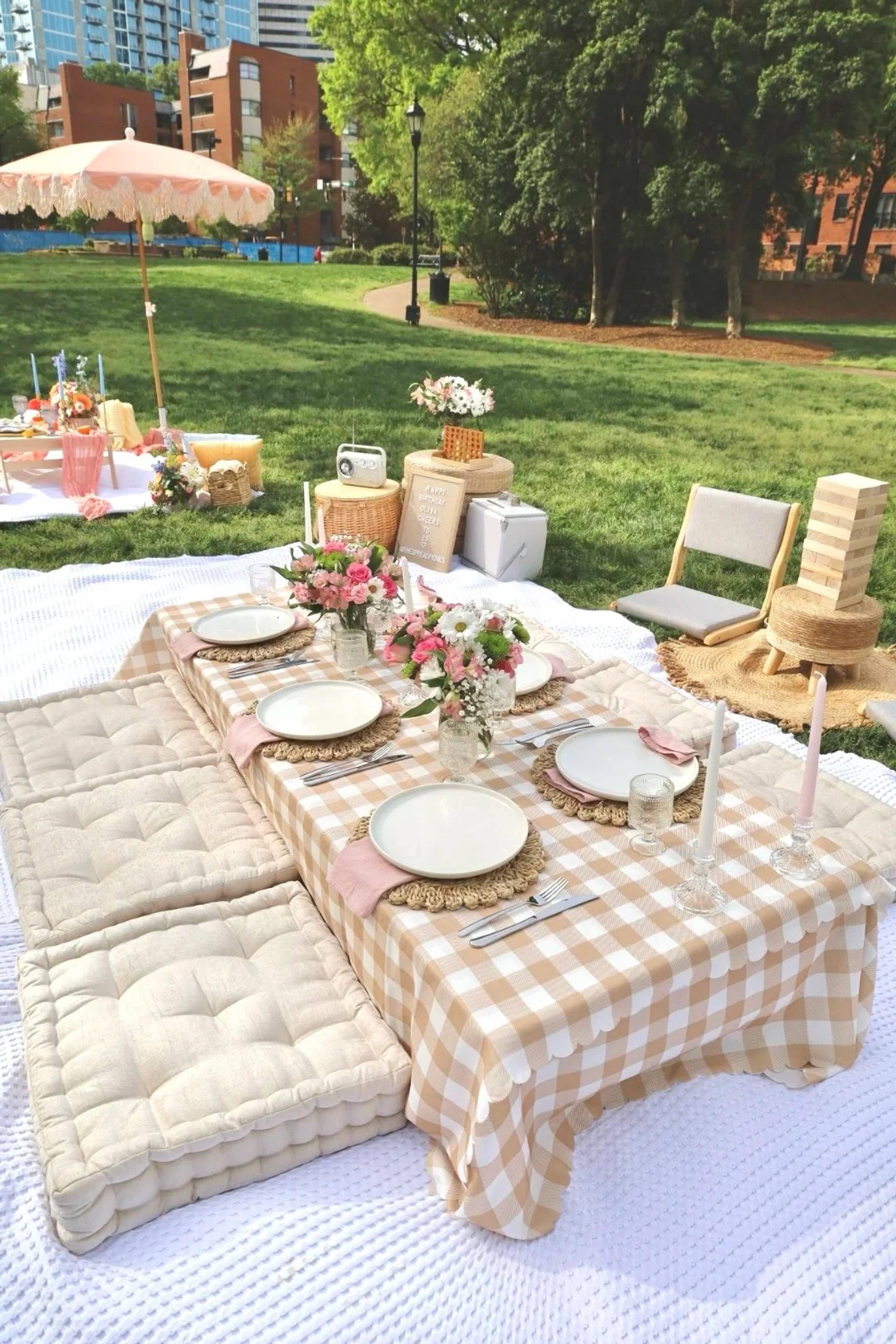 A gingham-covered low table set for six with floor cushions, pink flowers and candles, with a letter board sign, cooler and giant Jenga behind it