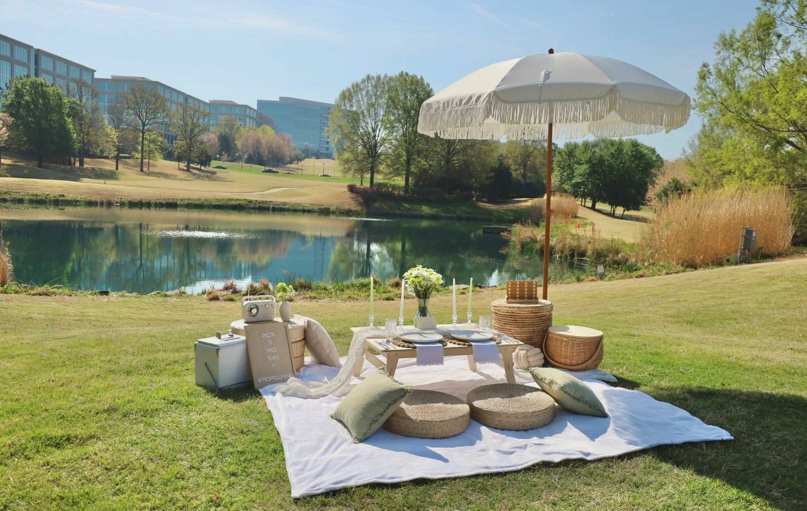 A picnic setup on a white blanket beside a lake, under a cream fringed umbrella