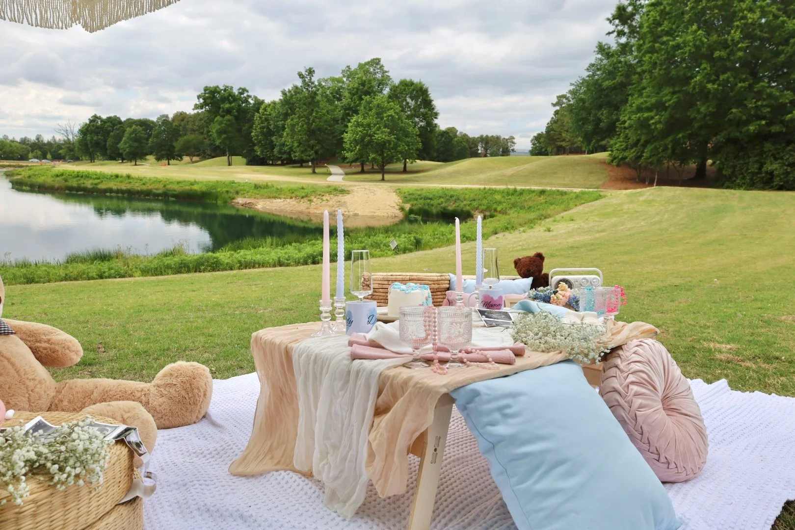 A picnic with floor cushions and a teddy bear on a lawn beside a pond, under a white umbrella