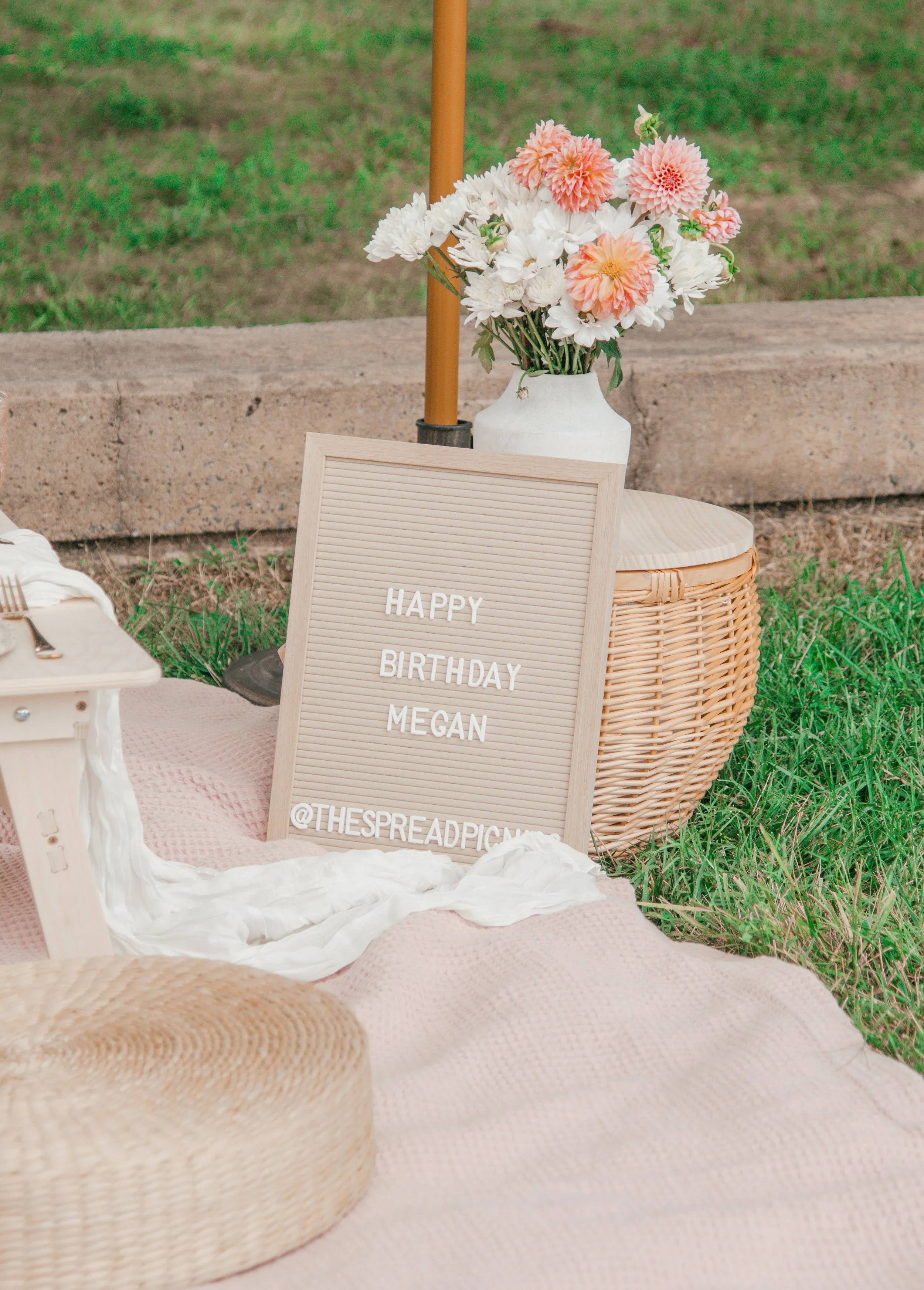 A Happy Birthday letter board beside a vase of peach and pink flowers on a picnic blanket
