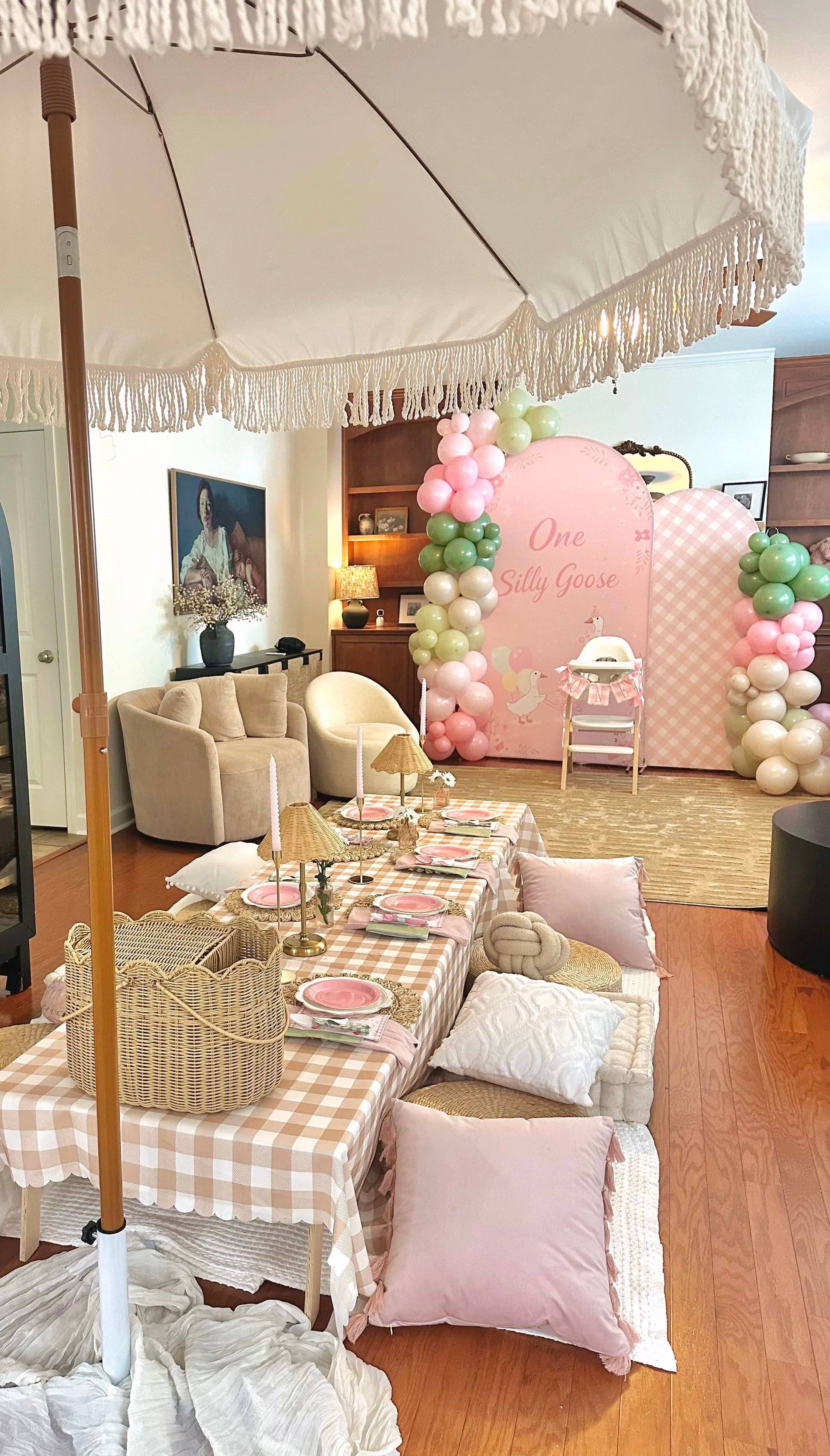 An indoor kids party: a long low gingham table with pink plates and cushions under a cream fringed umbrella, with a One Silly Goose balloon arch behind