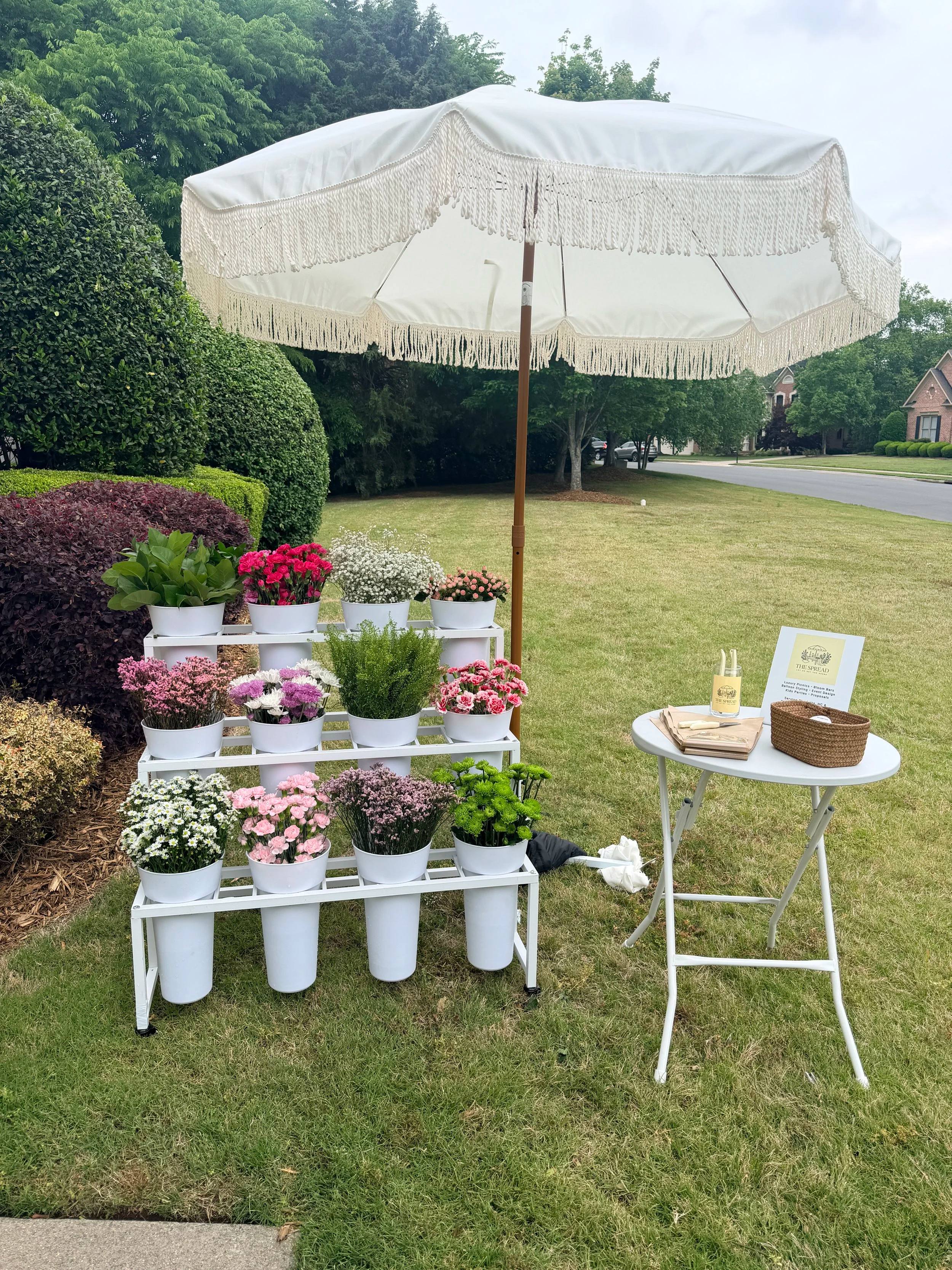 A white tiered Bloom Bar stand of flower buckets under a fringed umbrella on a lawn
