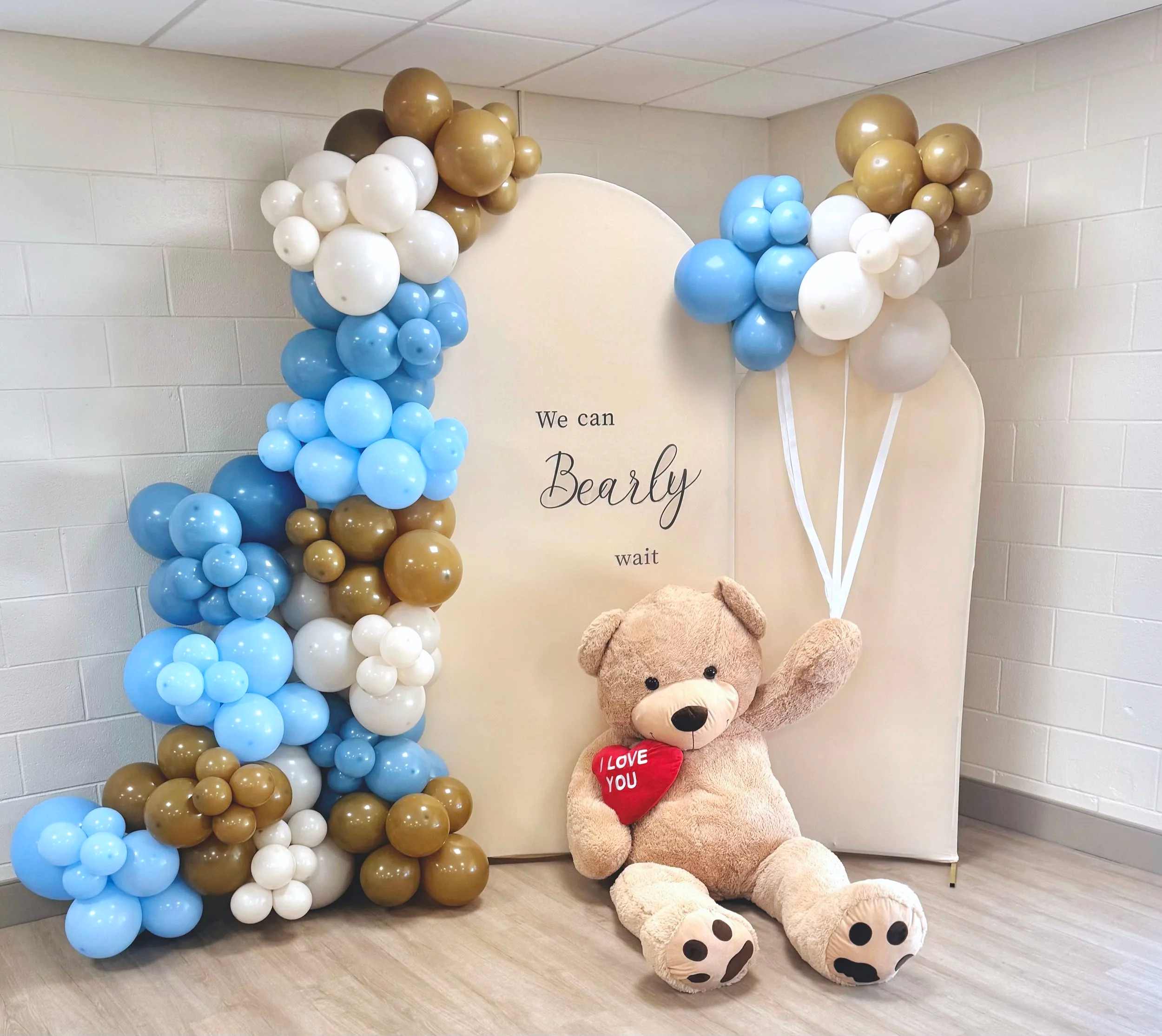 A We Can Bearly Wait baby shower backdrop with blue, tan and white balloon garlands and a giant teddy bear holding a red heart