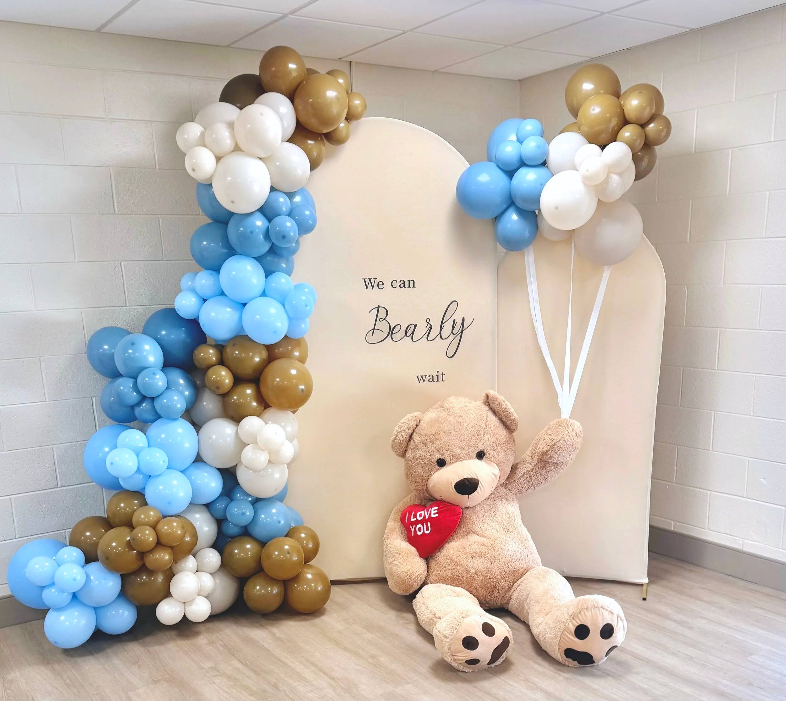 A We Can Bearly Wait baby shower backdrop with blue, tan and white balloon garlands and a giant teddy bear holding a red heart
