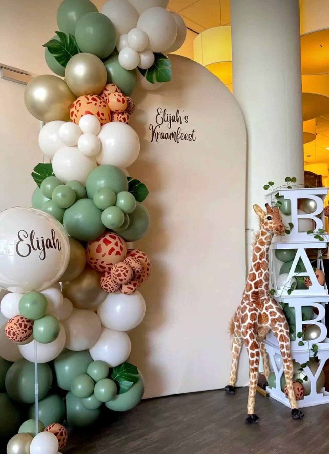 A safari baby shower backdrop with sage, cream and giraffe-print balloons, a toy giraffe and BABY blocks