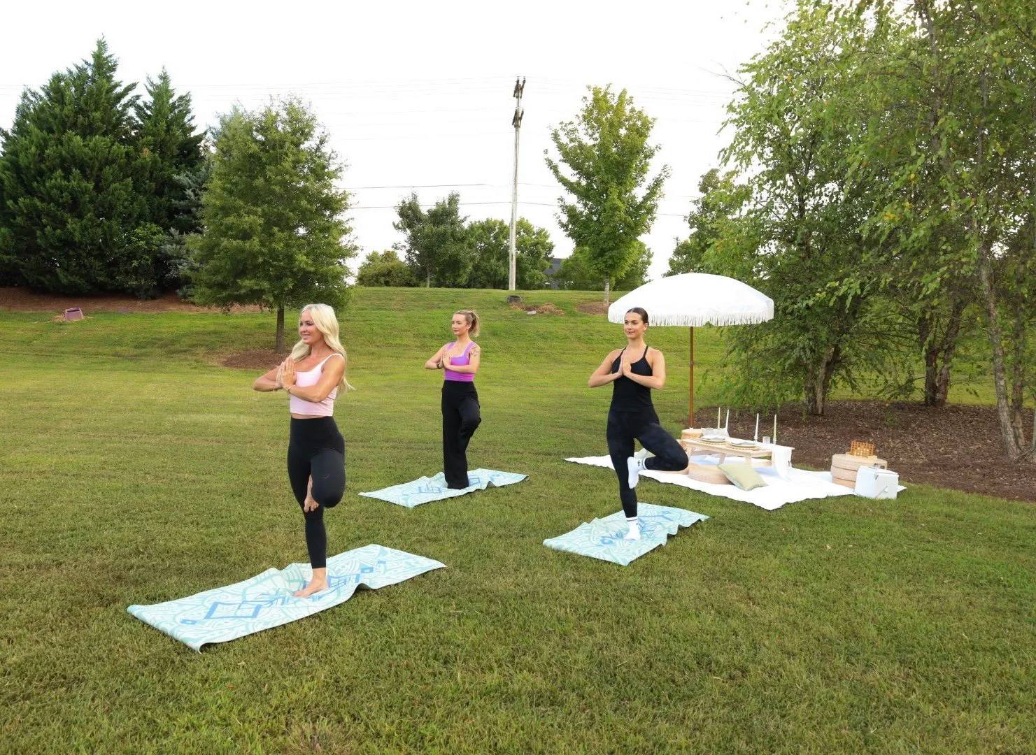 Three women doing yoga on mats beside a picnic on a lawn