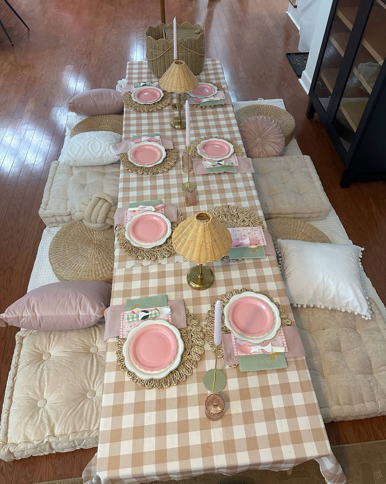 A long low gingham-covered kids party table indoors, set with pink scalloped plates, rattan lamps and pastel cushions on tufted mats