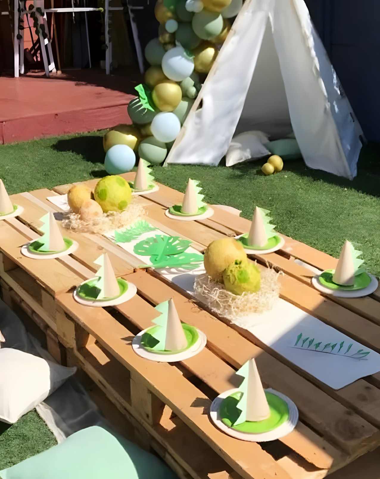 A kids party picnic on the grass: a low pallet table with green place settings and dino treat cones, white teepees and a balloon garland behind