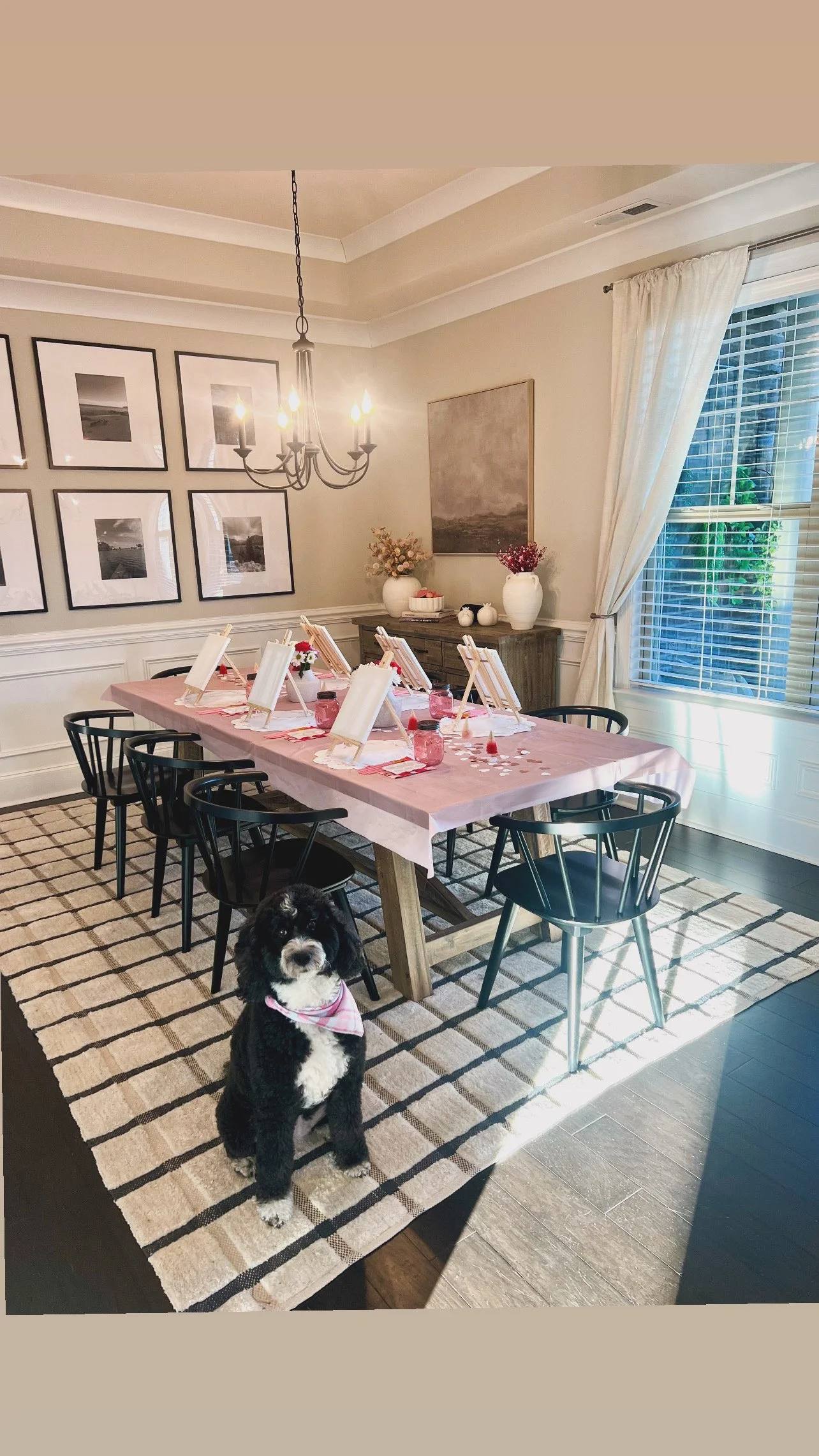 A dining room table dressed in pink and set for a party, with a black and white dog sitting on the rug in front