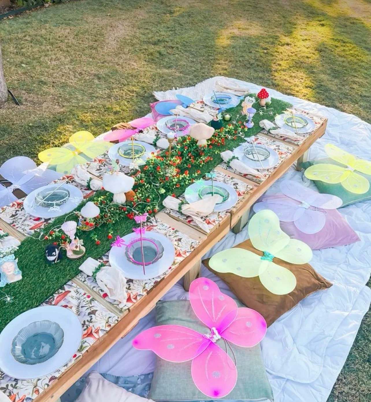 A fairy garden kids party table on a lawn: low pallet tables set with moss, toadstools, mini fairy figures, glass plates and pastel butterfly wings on the cushions