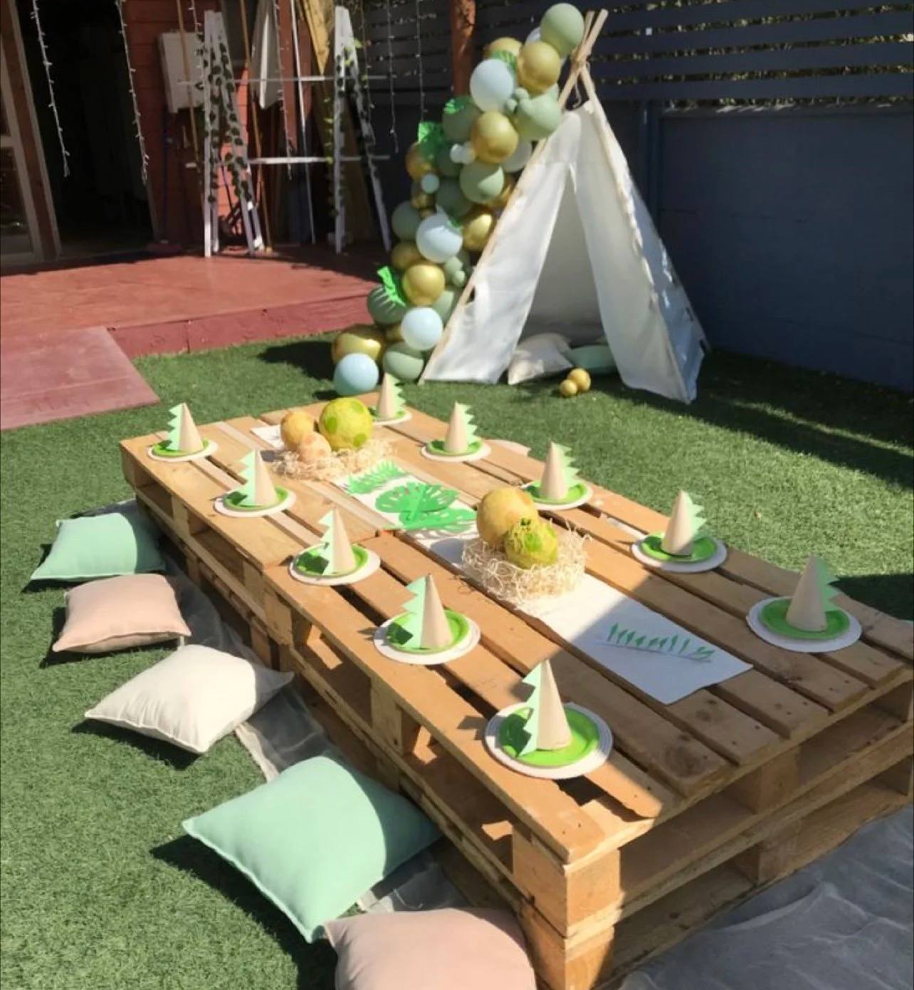 A kids party table built from low wooden pallets, set with green plates and party hats, floor cushions and a teepee with a green balloon garland
