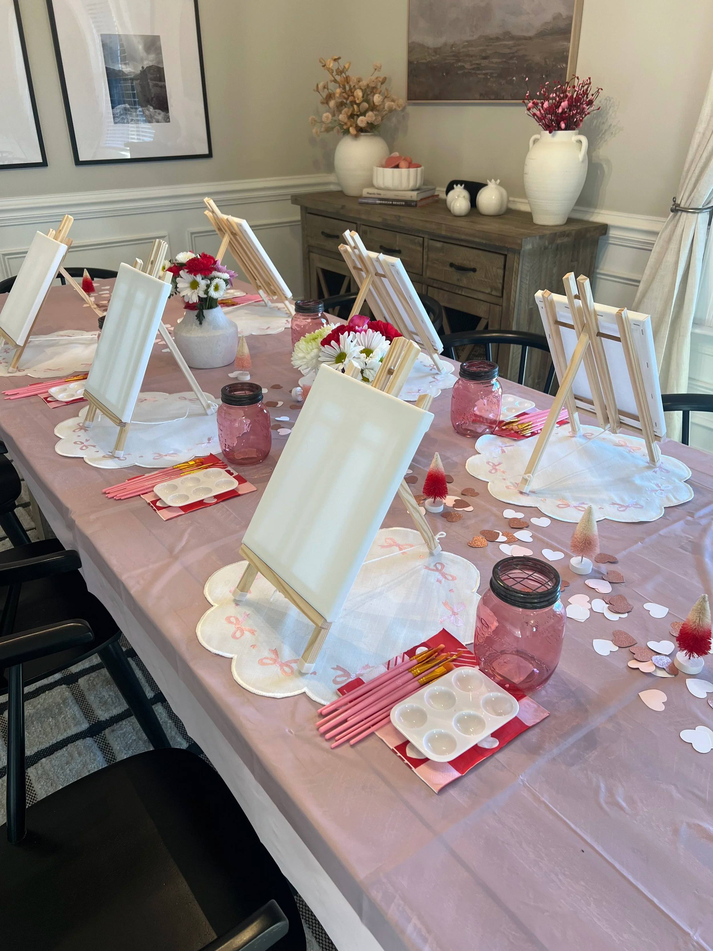 An indoor dining table set for a painting party with small easels, blank canvases, paint palettes and pink linens