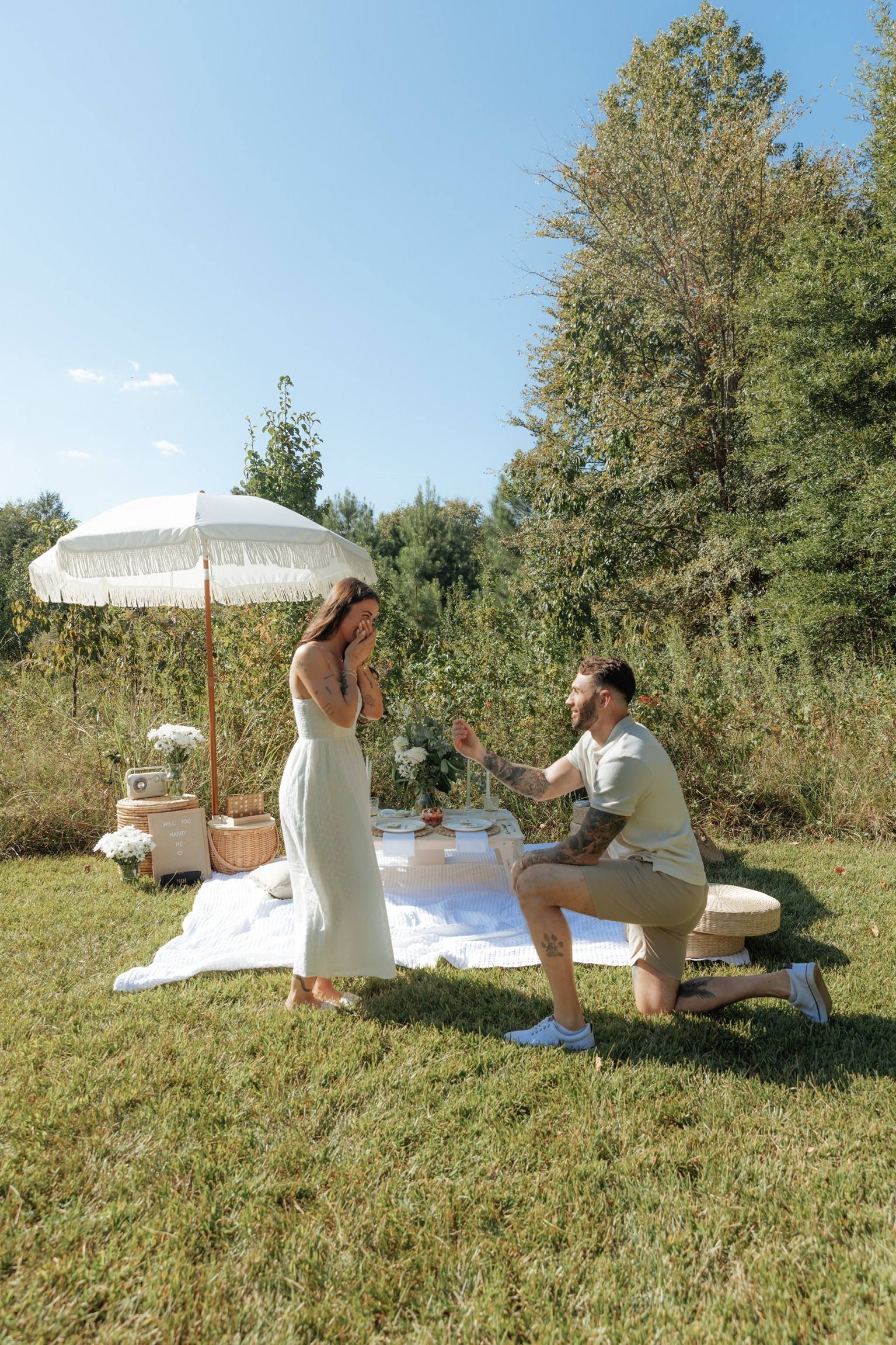 A proposal on a lawn: a man kneels in front of his partner beside a picnic under a white fringed umbrella