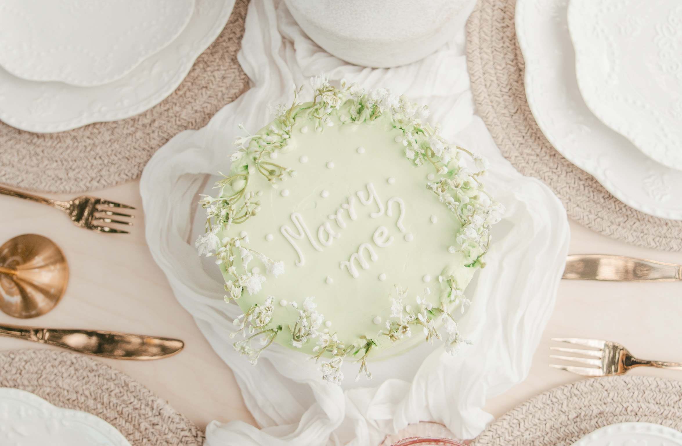 A pale green cake piped with Marry me? and ringed with baby's breath, seen from above between scalloped plates, gold cutlery and a pink glass