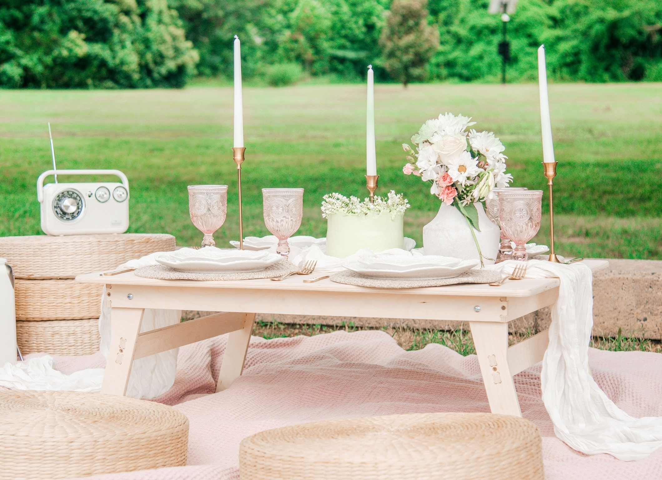 A low wooden picnic table on a pink blanket, set with a small cake, taper candles, pink glasses, a radio and a white runner, with wicker cushions on a lawn