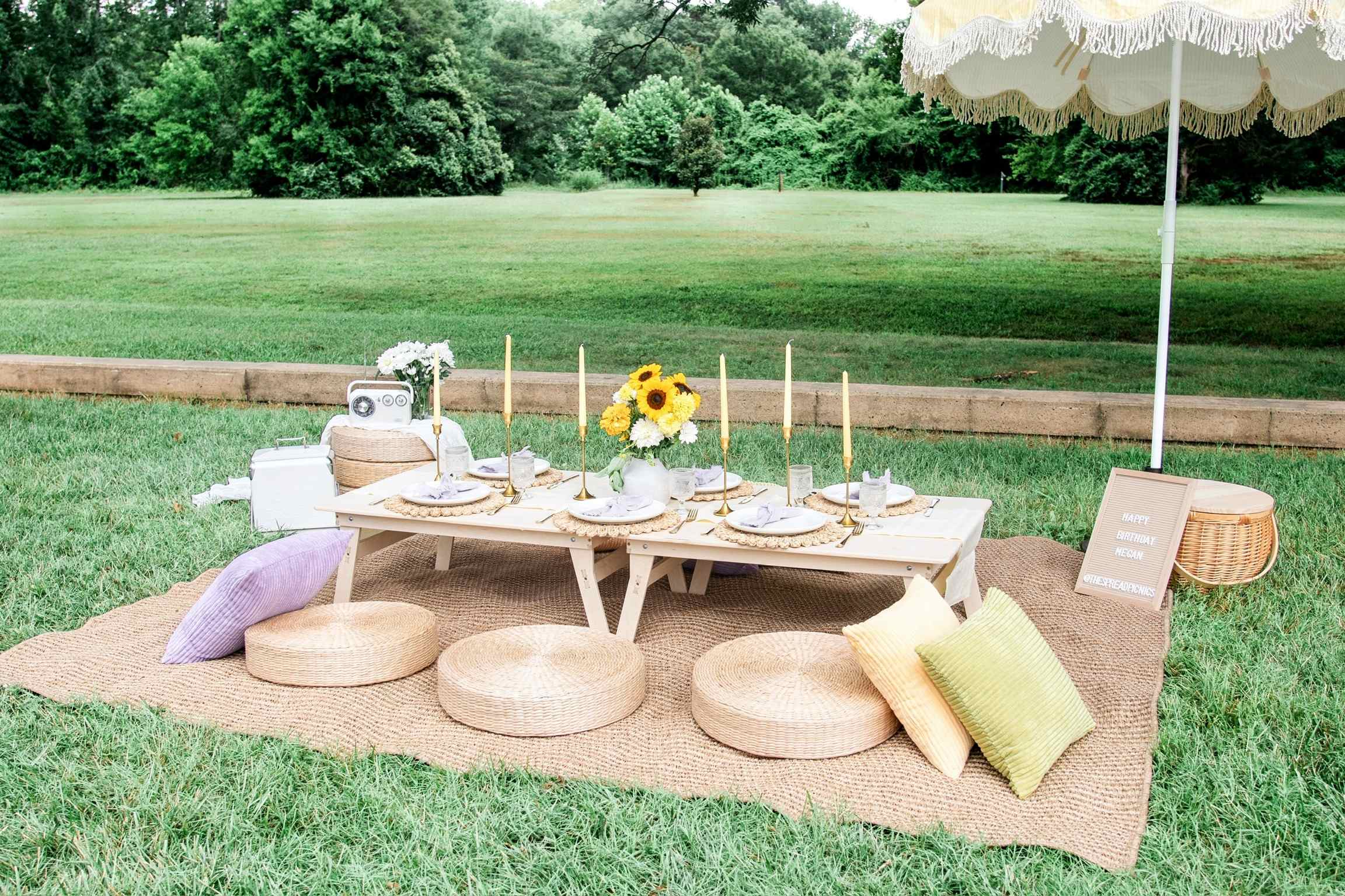 A low white picnic table set with sunflowers, gold candles and scalloped plates, with wicker and pastel cushions on a jute rug under a yellow fringed umbrella on a lawn